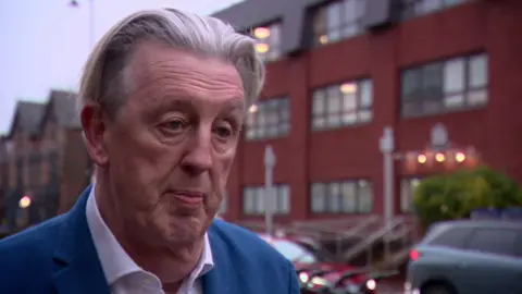 Paul Kohler, in a blue suit, stands in front of Wimbledon police station 