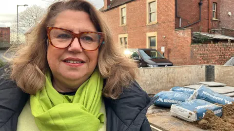 Helen Kowalski stands next to a builder's truck on a residential street. She is wearing a black jacket, green top and scarf and has thick-rimmed brown spectacles on. The truck behind her is filled with bags of cement.