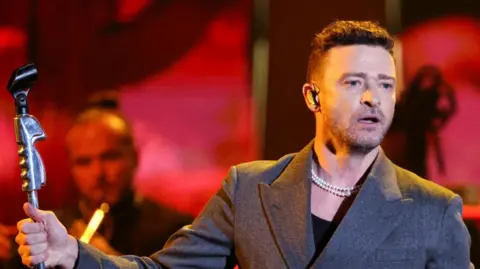 Justin Timberlake, a man in his mid-40s, stands on a stage holding a microphone stand. He is wearing a grey suit jacket and a necklace made of pearls. He has short brown hair and brown stubble and has a neutral expression. Another man playing a trumpet is in the background. The background is red.