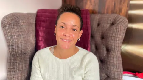 A woman looking at the camera and smiling. She is wearing a cream jumper while sitting in a brown tartan chair.