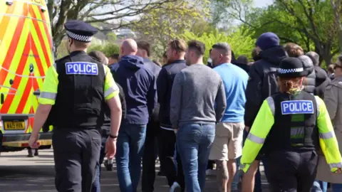 Two Devon and Cornwall Police officers walking with a number of football supporters outside Home Park Stadium in Plymouth. They all have their backs to the camera. There is a police van parked in the distance along with a number of trees. 