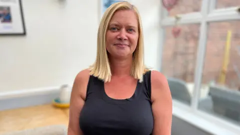Stuart Woodward/BBC A blonde woman with a black top on standing in a room and smiling for the camera. Behind her is a sunlit room with white walls.