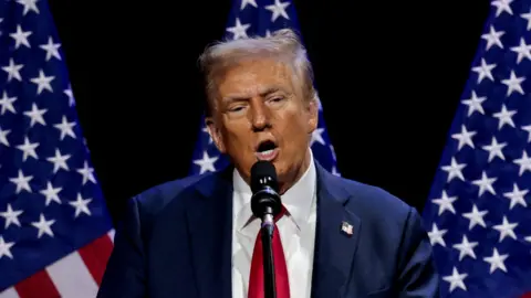 Reuters An image of Donald Trump, a man with short light hair wearing a blue suit, white shirt and red tie, speaking into a microphone standing in front of three US flags and a black background