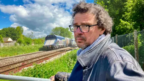 Simon Pope standing at the crossing where his mother was killed. He is wearing a dark leather jacket, a blue scarf, and has curly hair and glasses.