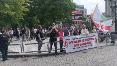 Protesters gathered ahead of an anti-immigration rally outside Belfast City Hall on Saturday. A small group of people are standing behind a banner which says "Stop uncontrolled immigration, we need to protect our children, we stand together." Two of the protesters are also holding up a flag which shows half of a Northern Ireland flag and half an Irish tricolour.
