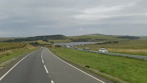 Google A30 road near the Jamaica Inn. Green fields surround the busy A-road. A lake can be seen in the background in between rolling hills. Cars are travelling on the road. 