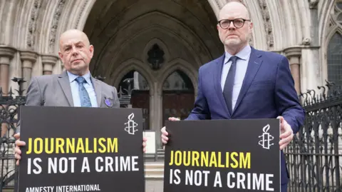 Barry on the left is bald, wearing a grey suit, blue shirt and blue tie.

Trevor on the right is taller, has a bit more hair than Barry and is wearing black round glasses. He is wearing a blue shirt and dark blue navy tie under a dark navy suit. 

They are both holding black signs that read 'Journalism is not a crime, Amnesty International' on them. 

Behind them, is the Royal Courts of Justice entrance, a grand, pillar shape with big windows. 