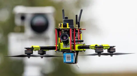 AFP via Getty Images A small drone with four rotors, three aerials and a camera on the front flies in front of a detection device.