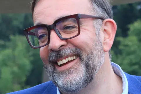 Kira Turnpenny Adam Nichols has a grey-flecked moustache and beard and is wearing large brown-rimmed glasses. He is looking away from the camera and laughing. He is wearing a blue jacket and a light blue shirt.