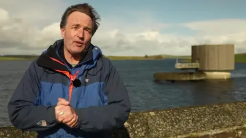 David Braine standing next to a reservoir. He is in a blue jacket looking at the camera. His hands are held together. It is a cloudy day.