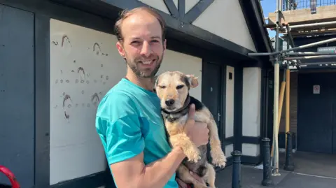 Andrew Gooder is wearing a bright turquoise shirt and is holding his terrier dog. Andrew has short brown hair and a beard.