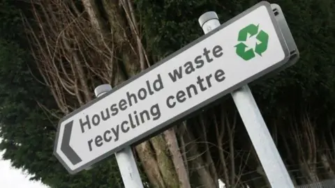 An arrowed sign with the words "Household waste recycling centre", alongside a green recycling logo.