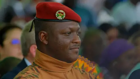 A head and shoulders shot of Burkina Faso's military leader Ibrahim Traore. He is wearing a red military beret and a camouflage roll-neck top.