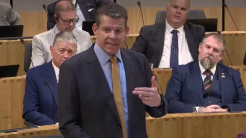Rhun ap Iorwerth stood up in the Senedd. He is wearing a black jacket, blue shirt and yellow tie. There are people sat behind him.