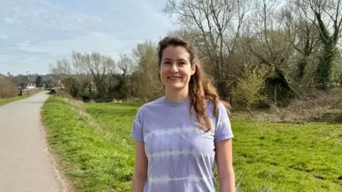 Liz Churchill, a woman in a violet coloured t-shirt stood on a path next to a park with lots of trees and grass around.