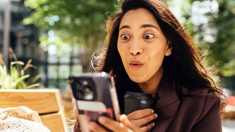 woman with long dark hair looks at her phone, with an expression of joy on her face.