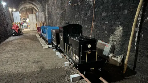 A miniature railway on small tracks in an arch-shaped tunnel with dark brick walls. The front of the train is back with silver railings round the side.