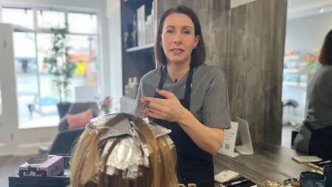 BBC Marie Hall wearing a grey t-shirt and black apron is standing in front of a woman who has her hair sectioned in to foils to be coloured. 