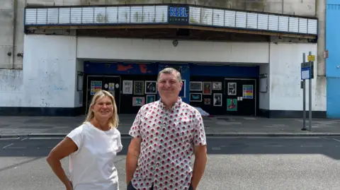 Hattie McCann/BBC Caroline Blacker and Karl Parsons are standing across the road from the front door of the old cinema. There are small posters plastering black boarding in front of the doors. The marquee, which is the display board where removable letters spelt out the names of the films, is just above the door. The words 'Reel cinema' are in the centre of it. Caroline has blonde hair and is wearing a white short-sleeved top and Karl has a white short-sleeved shirt with a red design on it.