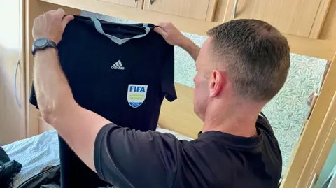 Ant Saddington/BBC A side profile of Stuart Burt holding up a black Adidas branded Fifa refereeing shirt. The shirt features a crest with the Fifa emblem and states 'assistant referee'. He is holding it above an open suitcase on a bed.