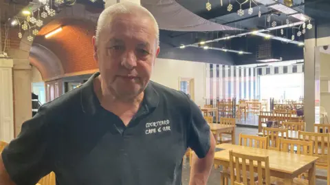 Simon Thake/BBC A man with shaved grey hair stands with his hands on his hips in an empty cafe