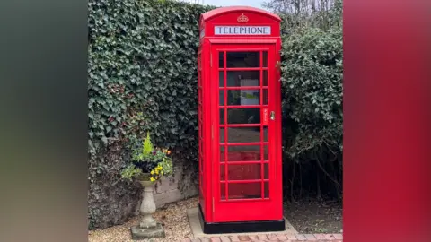 Greg Horsford A red phone box standing in the corner of a garden against an ivy clad wall. The phone box looks in pristine condition after being refurbished. 