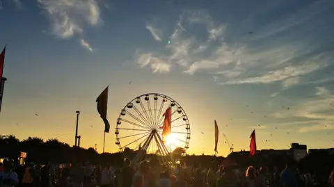 The sun slowly setting over a music festival crowd. There is a large ferris wheel and flags