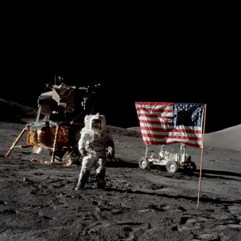 NASA Astronaut Harrison Schmitt in a white spacesuit stands on the surface of the Moon. Behind him is the lunar module with its gold panels and a lunar rover. In the foreground a US flag planted in the grey lunar dust. Behind the lunar terrain is a completely black sky.