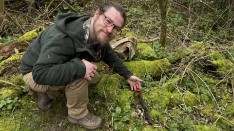 A man sits on a forest floor on a mossy log, wearing a green jumper, checked shirt and brown trousers. He is holding a red cap mushroom, which he found at Stanwick lakes nature reserve 