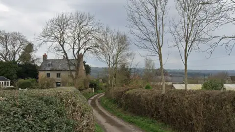 Google Maps A narrow country lane with tall hedges either side of it runs past a large three-storey old house.