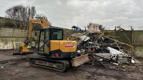 Phil Harrison/BBC A yellow construction vehicle with a grabber arm on a muddy plot of land. It is next to a large pile of scrap metal, where part of a sign reading Dymchurch Amusement Park can be seen.