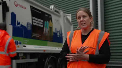 Rachel Jay of Veolia standing in front of a waste disposal lorry at the depot. She is wearing an orange high-viz jacket with the company logo on it.  