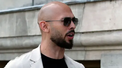 EPA Head and shoulders image of Andrew Tate standing outside a stone building. He is facing to the right, has a bald head and black short beard and is wearing sunglasses a black t-shirt and cream jacket