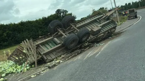 An overturned trailer with lots of cabbages strewn across the pavement at the side of the roundabout. In the background, a tractor can be seen