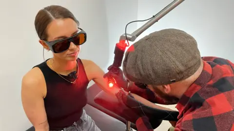 BBC A man wearing a tweed flat cap and a red and black checked shirt, is shown using a laser device to remove a tattoo on a woman's upper arm. The woman is wearing dark protective glasses and a black vest top. She is looking away from the tattoo as the man works on it.