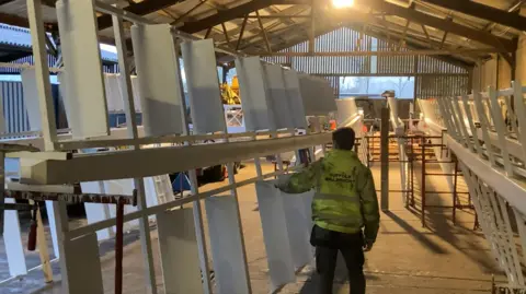 Luke Deal/BBC Mr Whiting walks through his warehouse and reaches out to a large white windmill sail. He has his back to the camera and he wears a florescent green hi-vis coat.