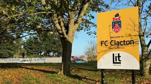 A yellow sign that says "welcome to FC Clacton" placed on grass in front of trees. In the background a wall that says "FC Clacton" can be seen, presumably a wall surrounding the team's ground. It is a clear autumn day with a blue sky.