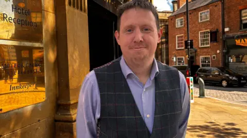 Simon Thake/BBC A man smartly dressed in a waistcoat and purple shirt smiles as he stands outside a stone building with a gold plaque on the wall