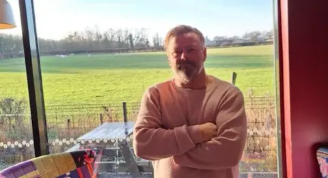 Local Democracy Reporting Service A man in a light coloured top stands with his arms folded in front of a large window. Behind him is a picnic table and fence and behind that is a field with sheep in and trees in the distance.