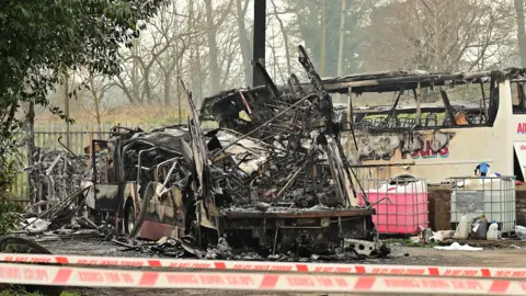 Pacemaker Two burnt out buses, one is black metal melted parts and another still has a partial boyd frame, it is a white bus with pink writing that says Allen's Tours. There is other debris at the buses and a police cordoned line with white and red tape - in red it says DO NOT CROSS and POLICE CORDON 