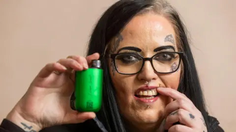 A woman holds a green vape up between her thumb and index finger beside her face while holding back her upper like with her other index finger. she has long straight black hair dark rimmed glasses, facial jewellry on her nose and lip and small tattoos on her cheeks and forehead 
