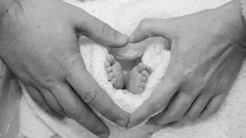 Martina and Ryan Tierney A black a white image of a baby's feet with hands around them pointing their fingers together to shape a heart. The baby's feet is pictured in a white blanket. 