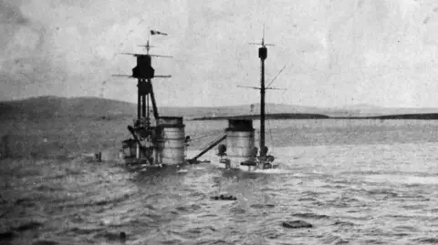 Photo 12/Getty Images The funnels and masts of a sunken ship protrude from the water with the islands of Orkney in the background in this grainy black and white photo.