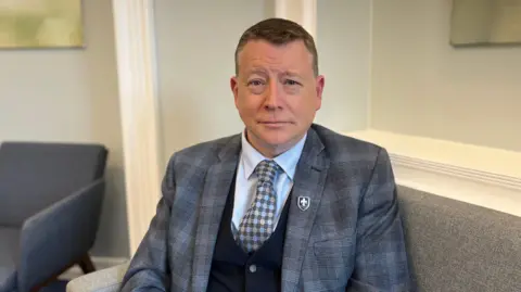Matthew Burke is pictured sitting in an office at Beaulieu school in Jersey. He is wearing a plaid blazer, waistcoat and spotty tie. He has short brown hair and is smiling. 