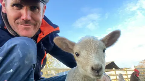 Mark Rutherford A man in a flat cap and outdoor working clothes, knelt down smiling into the camera. Next to him is a sheep, and behind is a farm.