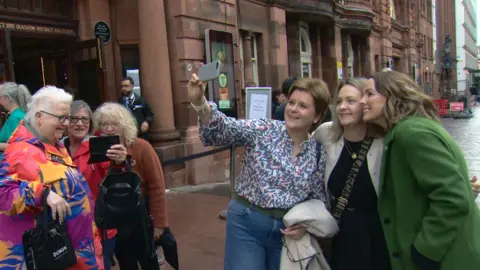 Nicola Sturgeon, dressed in jeans and a patterned blouse, poses for selfies, with Val McDermid also seen, dressed in a brightly coloured coat.