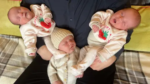 Three babies are being held by a man in a blue shirt. The babies are wearing white clothes and are sleeping.