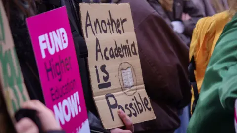 UCU A close up of two signs on a picket line held by university staff on strike over job losses. On the left a bright pink printed sign reads "Fund Higher Education Now". The other sign is handwritten on cardboard and says "Another Academia is possible".