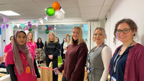 Eleven women stand in a health room. They are cutting a ribbon and there are balloons behind them.