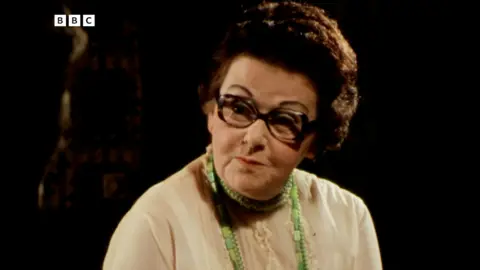 Peggy looking to the side in a dark room. She's got pursed lips like she's just about to open her mouth.